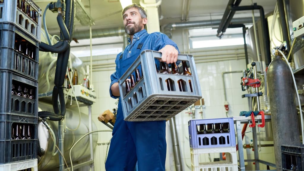 Man in blue work overalls carrying a crate of empty glass bottles inside an industrial facility with machinery and equipment, illustrating part of the process at a site operating under a liquor manufacturer license.